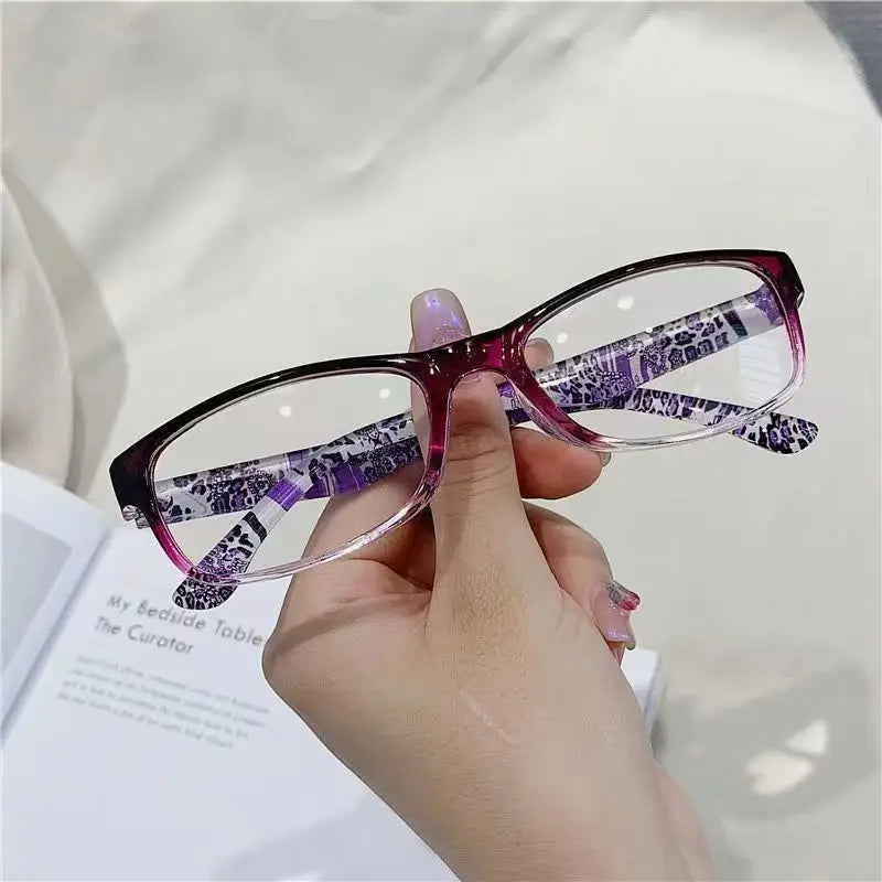 A hand holds a pair of purple-framed reading glasses with a patterned lens against a light background.