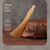 A clear, elongated wooden tool used for facial scraping therapy, tapping, and plucking tendon massage on a dark wood surface.
