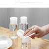 A hand is squeezing a white substance from a clear refillable push-down pump dispenser bottle onto a wooden table.