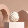 A white deep tissue massage ball sits on a light gray pedestal against a soft pink background.