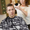 A man wearing a black patterned cape with various barber tools and styling accessories, being attended to by a woman.