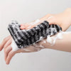 Hands are applying a black exfoliating loofah to the skin, creating white foam.