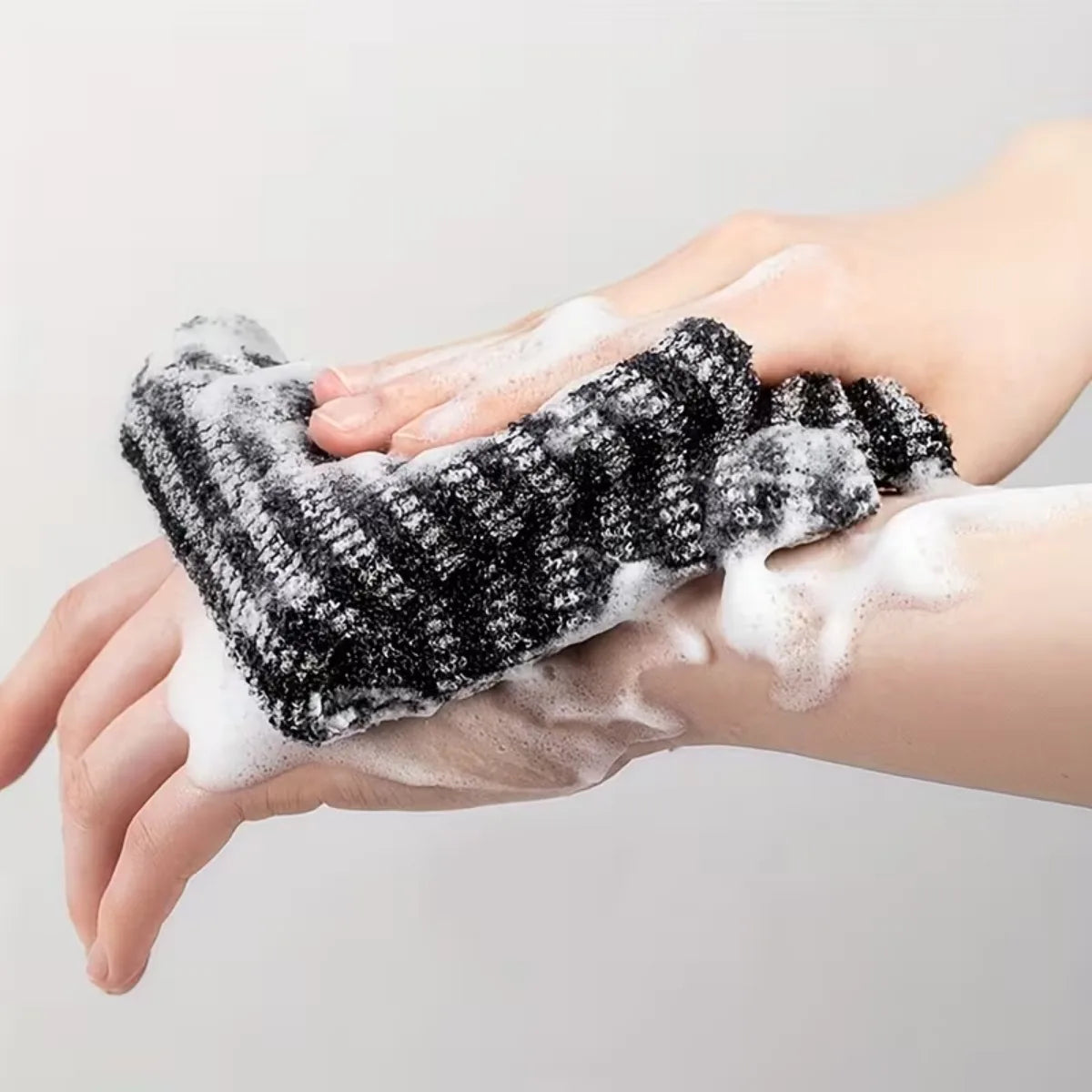 Hands are applying a black exfoliating loofah to the skin, creating white foam.