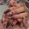 A close-up of a hand displaying 24-piece almond press-on nails featuring floral and butterfly designs.
