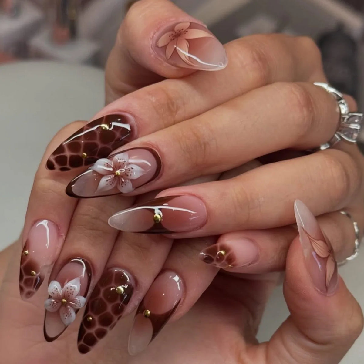 A close-up of a hand displaying 24-piece almond press-on nails featuring floral and butterfly designs.