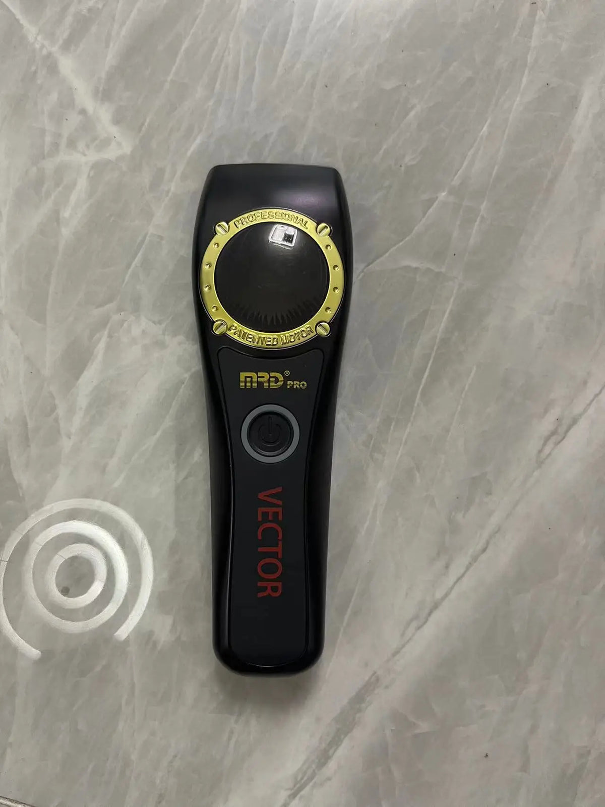 A black professional electric hair trimmer with a silver and gold accent ring is shown on a light marble surface.