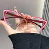 A hand holds a pair of pink cat eye reading glasses with dark red frames against a light gray background.
