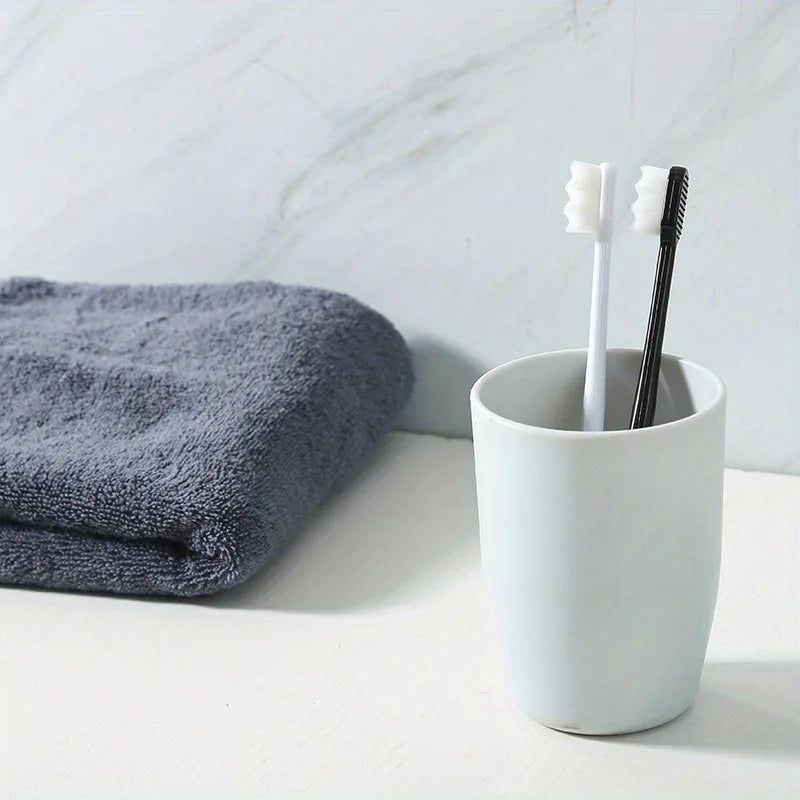 A white ceramic cup holds a toothbrush and a toothbrush holder next to a gray towel on a white marble surface.