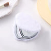 A heart-shaped foldable travel makeup mirror with a silver frame and white fluffy material is shown on a white surface.