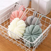 Three soft exfoliating bath bubble balls in pink, beige, and green are displayed in a white mesh basket.