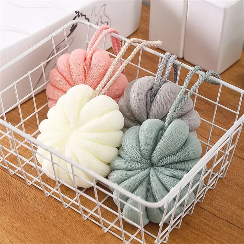 Three soft exfoliating bath bubble balls in pink, beige, and green are displayed in a white mesh basket.