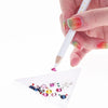 A hand holds a white pencil with a brush tip, applying colorful rhinestones onto a clear surface for nail art.