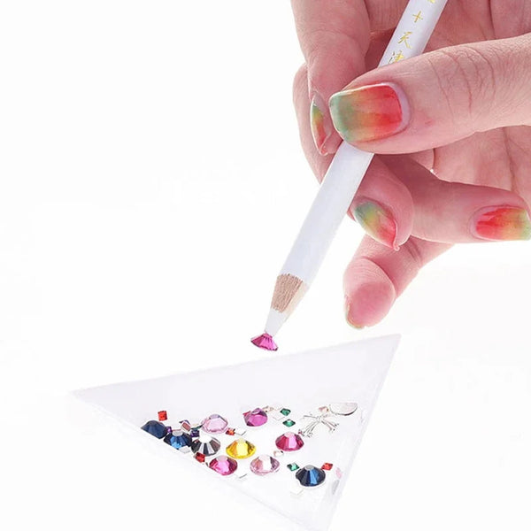 A hand holds a white pencil with a brush tip, applying colorful rhinestones onto a clear surface for nail art.