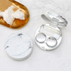 A round marble pattern contact lens storage box with a silver lid and a gold dish containing white cosmetic product.