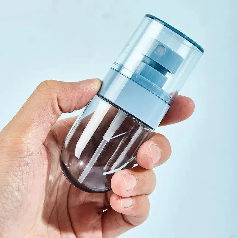 A hand holds a clear, portable refillable fine mist spray bottle with a blue cap and nozzle.