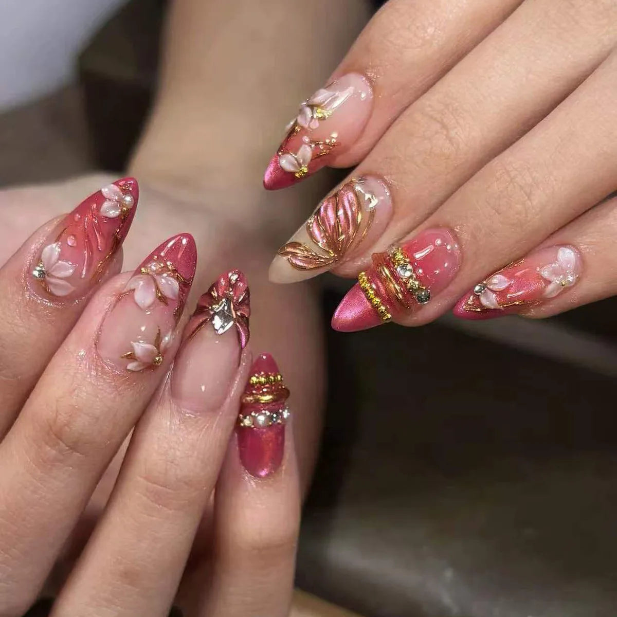 A close-up of a hand displaying 24-piece almond press-on nails featuring pink and gold floral and butterfly designs.
