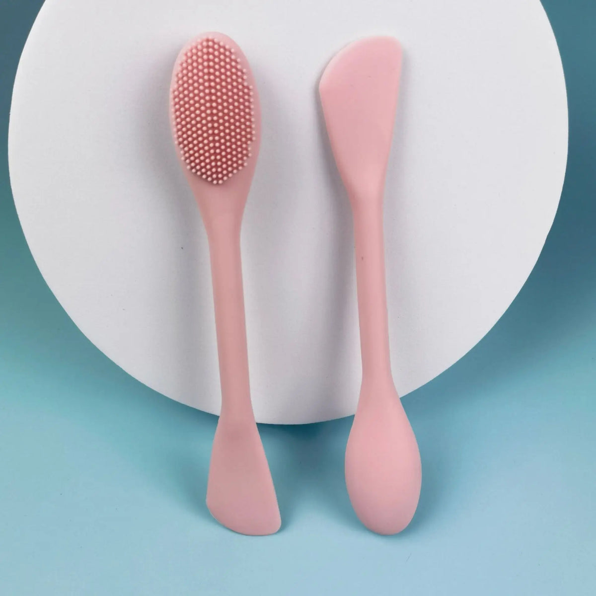 A pink double head silicone face cleansing brush resting on a white circular background.