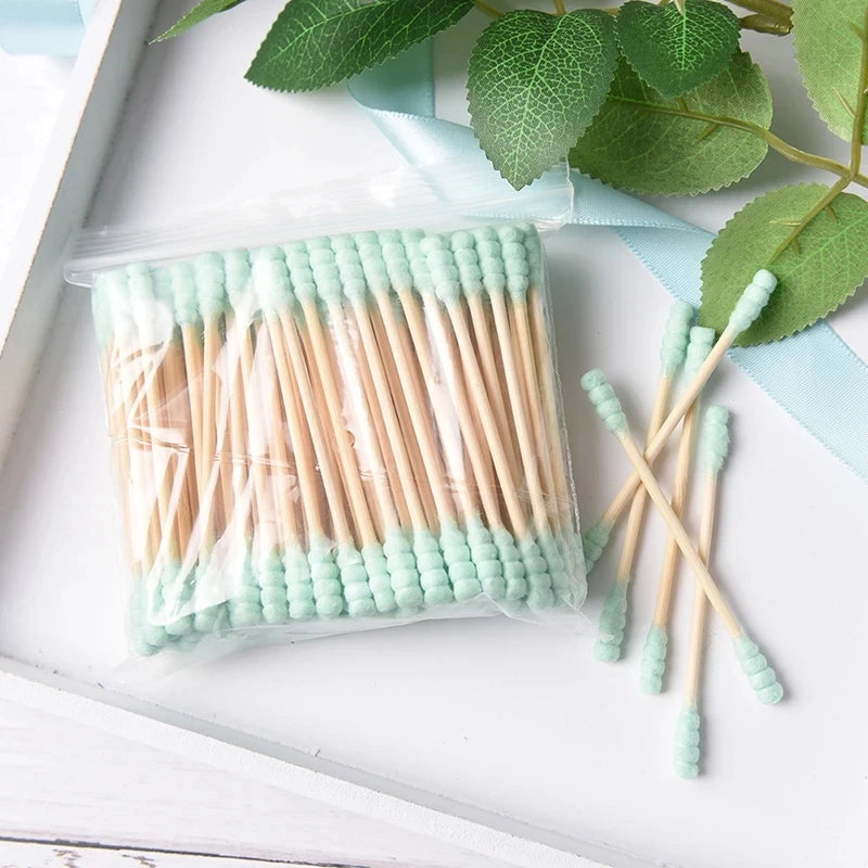 Bâtonnets de cotons en bambou avec des pointes vert clair et des tiges en bois naturel, posés sur une surface blanche
