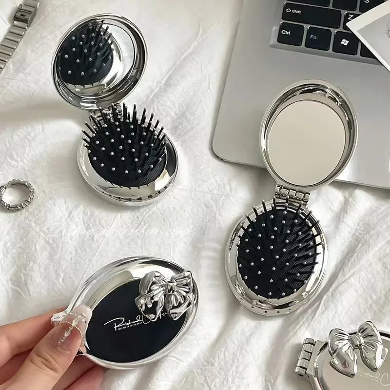 A silver folding massage comb with black bristles and a mirror is shown on a white surface.