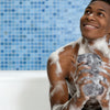 A young man is using an African net exfoliating loofah sponge in a bathtub with blue tiled walls.