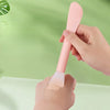 A hand is using a pink silicone facial mask brush with an integrated scraper to apply product onto the skin.