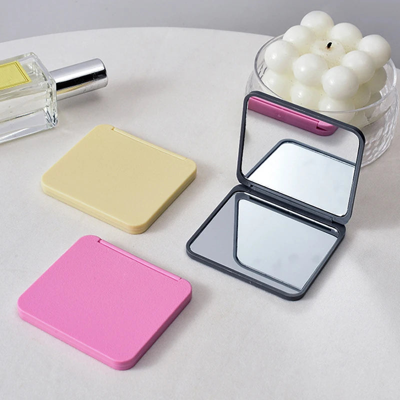 A set of double-sided portable makeup mirrors in pink and yellow, next to a glass candle holder.