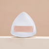 A white triangular cosmetic foundation puff brush with a pink band resting on a light beige surface.