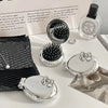 A silver folding massage comb and mirror are displayed on a white surface next to some fabric.