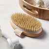 Un cepillo exfoliante de cuerpo con cerdas naturales reposando sobre un plato de madera con una almohadilla exfoliante blanca
