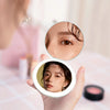 A hand holds a circular LED makeup mirror showing a woman's eye with luminous glitter effect.