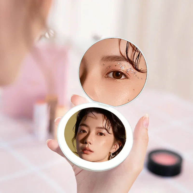 A hand holds a circular LED makeup mirror showing a woman's eye with luminous glitter effect.