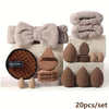 A set of professional makeup brushes and sponge in various shades of beige and brown stacked on a white surface.