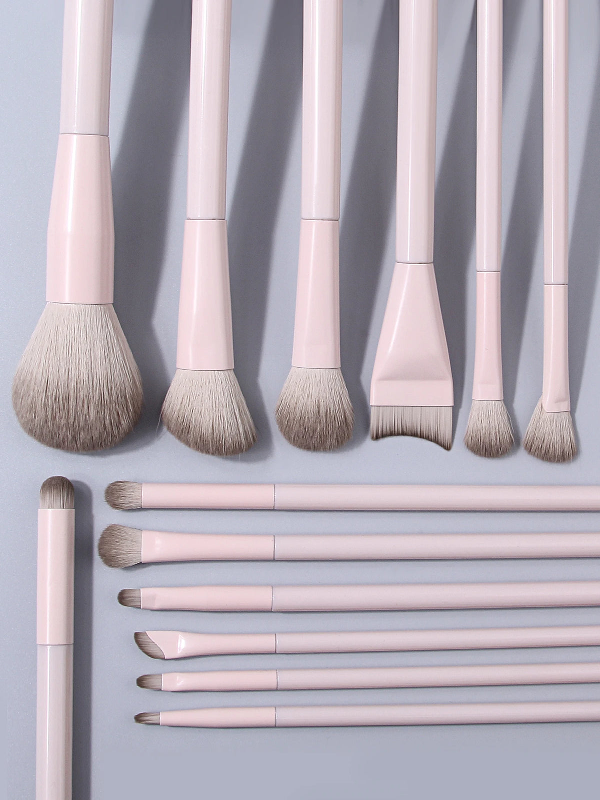 A set of thirteen professional makeup brushes in a pale pink color displayed vertically.