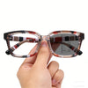 A hand holds a pair of brown and white patterned photochromic reading glasses.