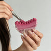 A hand is using a pink silicone gua sha tool with pink facial glitter on a light gray background.