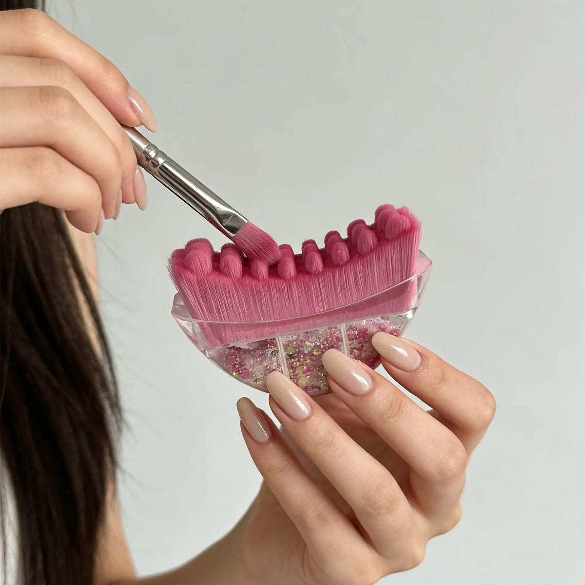 A hand is using a pink silicone gua sha tool with pink facial glitter on a light gray background.