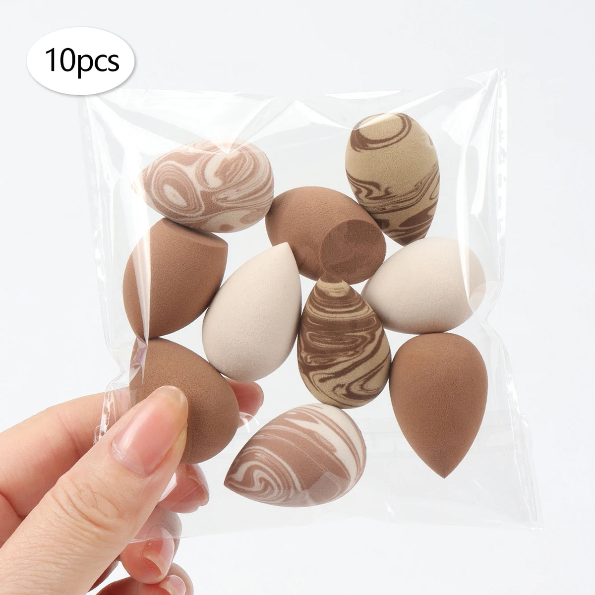 A hand holds a clear plastic bag containing 10 reusable expandable makeup sponges in various natural and earthy tones.