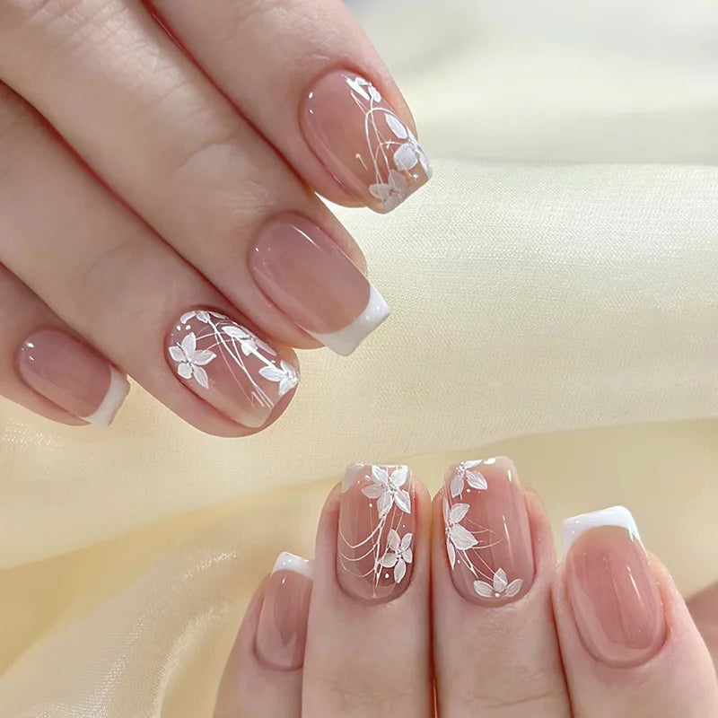 A close-up of a hand displaying nude pink press-on nails decorated with white floral accents and small 3D charms.