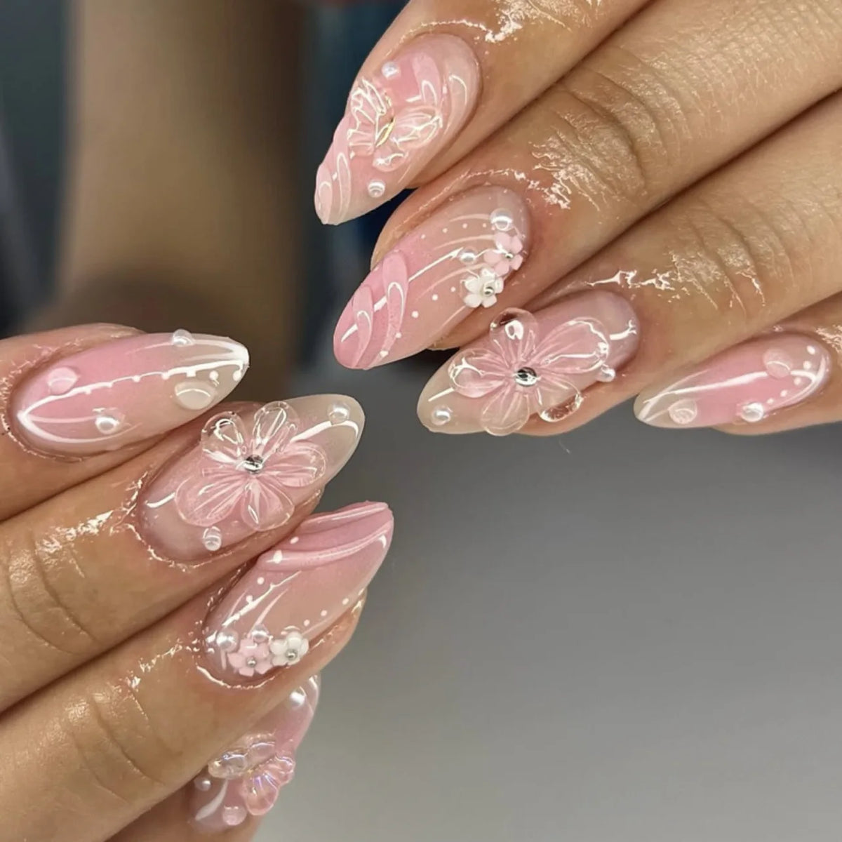 Close-up of a hand displaying 24-piece almond press-on nails featuring delicate pink floral and butterfly designs.