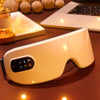 A white smart eye massager device resting on a wooden surface with soft ambient lighting.