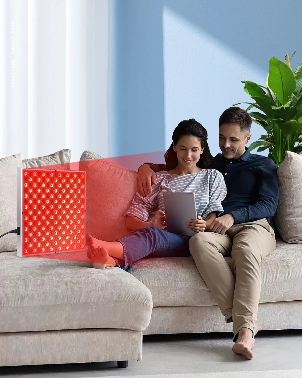 A woman is sitting on a sofa reading a book while using a red light therapy panel for skin and pain relief.