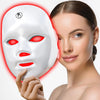 A woman is applying a white LED facial mask to her face, which emits a red glow.