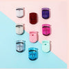 A collection of mini folding eyelash curlers in various colors including white, purple, red, pink, and teal, arranged on a li
