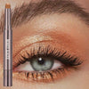 A cosmetic product featuring a long-lasting eyeshadow stick with a double brush placed next to an eye applying warm-toned eye