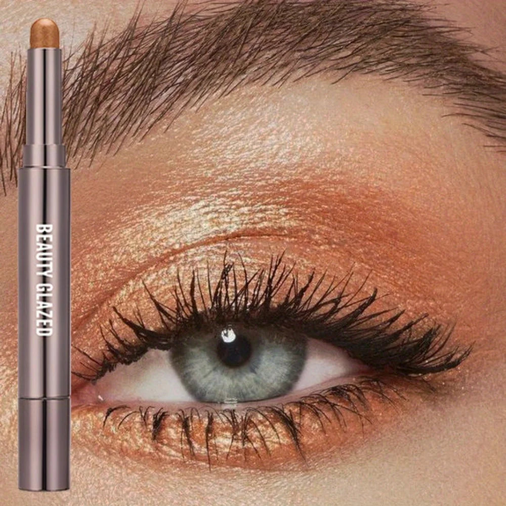 A cosmetic product featuring a long-lasting eyeshadow stick with a double brush placed next to an eye applying warm-toned eye