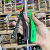 A hand holds a black folding shaving razor and a green folding shaving razor against a blurred background of other grooming p
