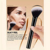 A close-up of a black makeup brush with a fluffy ferrule and a handle, shown next to a woman's face.