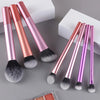 A set of seven professional makeup brushes in various shades of pink and purple displayed on a gray surface.