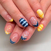 Close-up of a hand displaying reusable press-on nails with various blue and yellow stripes and polka dots on an almond shape.
