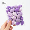 A hand holds a clear plastic bag containing 30 reusable expandable makeup sponges in various shades of purple and lilac.